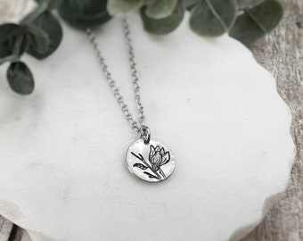 Puede incluir: Un collar de tono plateado con un pequeño colgante redondo de metal martillado. El colgante está grabado con un dibujo detallado en líneas negras de una flor con hojas en un tallo. El colgante cuelga de una delicada cadena plateada.