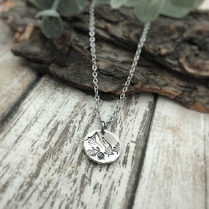 May include: A silver-toned circular pendant necklace featuring an engraved design of a mountain range, a bicycle, and pine trees. The pendant hangs from a delicate silver-toned metal chain, showcasing a detailed outdoor-themed graphic.