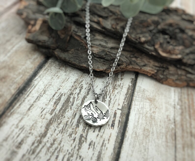 May include: A silver-toned circular pendant necklace featuring an engraved design of a mountain range, a bicycle, and pine trees. The pendant hangs from a delicate silver-toned metal chain, showcasing a detailed outdoor-themed graphic.