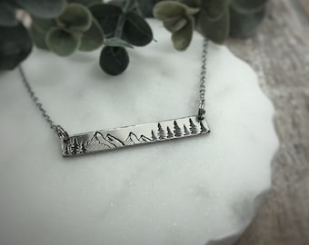 May include: A silver-toned horizontal bar necklace featuring an engraved landscape design of mountain peaks and evergreen trees. The rectangular metal bar is attached to a delicate silver-toned chain on both ends, creating a clean and simple accessory.