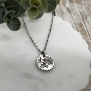 May include: A silver necklace with a round pendant featuring a mountain scene with a bicycle. The pendant has a hammered texture.