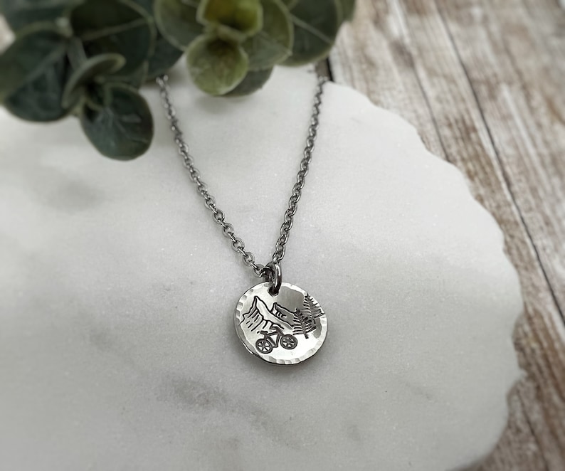 May include: A silver-toned metal necklace featuring a small, round, hammered pendant. The pendant is engraved with a detailed black graphic depicting a mountain range, a pine tree, and a bicycle. The pendant hangs from a delicate silver-toned chain.