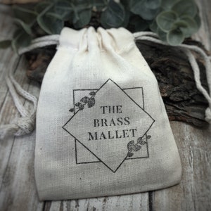 May include: A small, off-white drawstring pouch made of woven fabric. The front features a black printed design consisting of a square and diamond shape, with small leaf illustrations and the text THE BRASS MALLET centered in the middle.