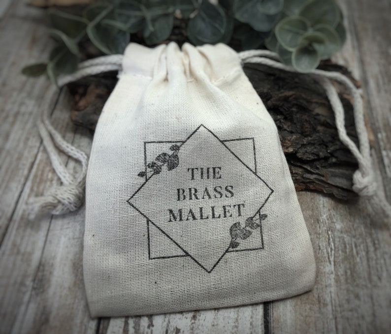 May include: A small, off-white drawstring pouch made of woven fabric. The front features a black printed design consisting of a square and diamond shape, with small leaf illustrations and the text THE BRASS MALLET centered in the middle.