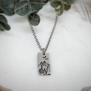 Könnte beinhalten: Eine silberne Halskette mit einem rechteckigen Anhänger, der eine gravierte Bergszene mit Bäumen und Vögeln im Flug zeigt.