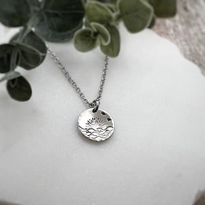 Puede incluir: Un collar de plata con un colgante redondo que representa un sol poniéndose sobre las olas y dos pequeños corazones.