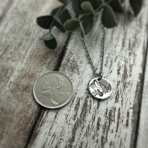 May include: A silver-toned circular pendant necklace featuring a stamped mountain, bicycle, and pine tree design. The pendant is shown next to a Canadian 25-cent coin for scale, which has a diameter of approximately 0.94 inches, on a thin metal chain.