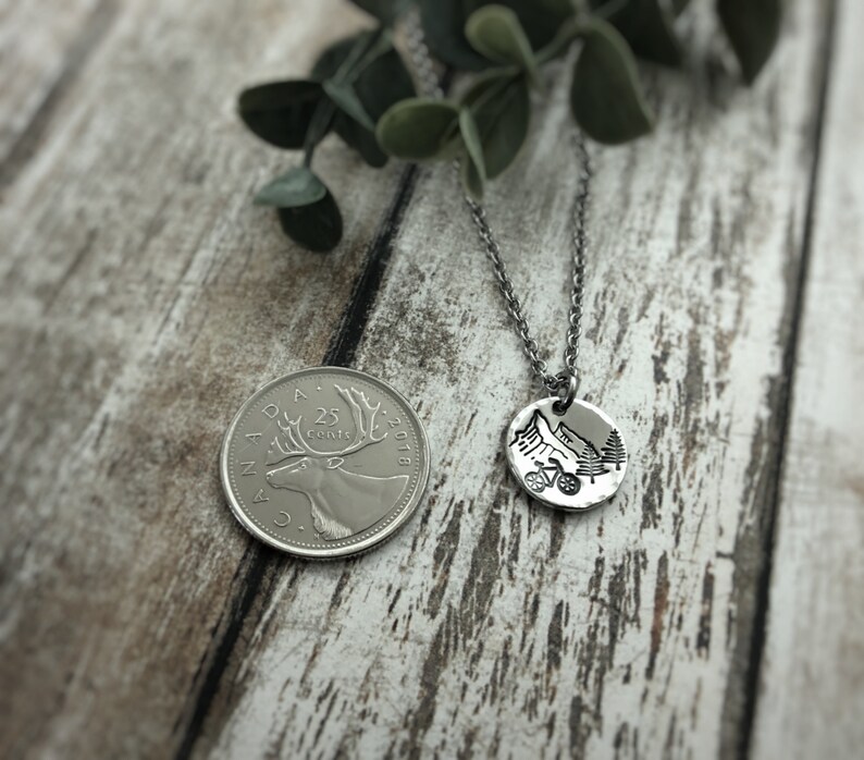 May include: A silver-toned circular pendant necklace featuring a stamped mountain, bicycle, and pine tree design. The pendant is shown next to a Canadian 25-cent coin for scale, which has a diameter of approximately 0.94 inches, on a thin metal chain.
