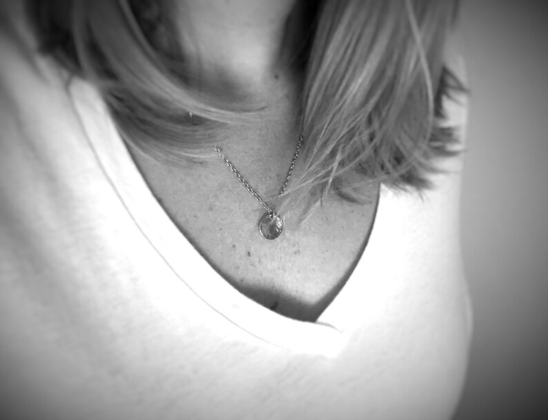 May include: A silver-toned necklace featuring a delicate chain and a small, circular pendant with a textured, metallic surface. The pendant hangs centrally from the chain, resting against a plain white v-neck shirt in this black and white image.