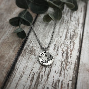 May include: A silver-toned circular pendant necklace featuring a stamped black graphic of a mountain range, a bicycle, and pine trees. The pendant hangs from a matching silver-toned metal chain link necklace, showcasing a hammered texture.