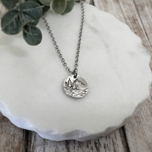 May include: A silver necklace with a round pendant featuring a hand-drawn design of a mountain range with trees and a lake. The pendant is textured and has a hammered finish.