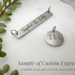 May include: A silver-toned rectangular bar pendant engraved with the text Sophie & Lily and a circular pendant engraved with the letters S & L. Both metal charms feature small jump rings attached to the top for hanging on a necklace chain.
