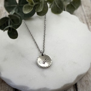 May include: A silver-toned metal necklace featuring a small, round, hammered-texture pendant. The pendant is engraved with a black infinity symbol intertwined with an arrow. The pendant hangs from a delicate silver-toned metal chain link necklace.