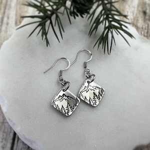 May include: A pair of silver earrings with a square-shaped charm featuring a mountain range and trees. The earrings are on a white surface.