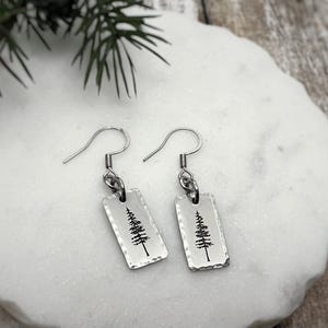 May include: Silver-coloured earrings with rectangular pendants, each with a hammered texture and a black tree design. The earrings are shown on a white, textured surface, with a sprig of greenery in the background. The earrings are a nature-themed accessory.
