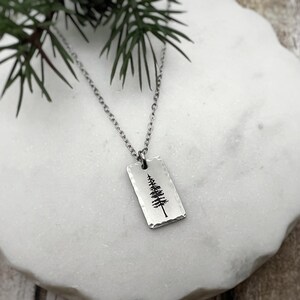 Puede incluir: Un collar plateado con un colgante rectangular. El colgante tiene una textura martillada y un diseño de árbol negro. El collar se muestra sobre una superficie blanca con efecto mármol, con follaje verde en el fondo.