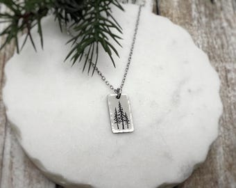May include: A silver-toned rectangular pendant necklace featuring a hammered texture and a black engraved graphic of three pine trees. The pendant hangs from a delicate silver-toned metal chain, showcasing a detailed forest-inspired design.
