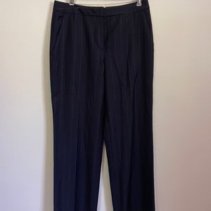 May include: Navy blue pinstripe trousers hanging on a wooden hanger. The pants have a straight leg cut and feature vertical stripes. The trousers are made of a smooth fabric and have pockets.
