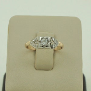 Puede incluir: Un anillo de compromiso vintage con un solo diamante redondo engastado en un bisel cuadrado. El anillo está hecho de oro amarillo y blanco y presenta un delicado halo de diamantes.