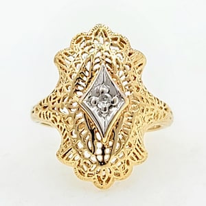 May include: A gold filigree ring with a diamond-shaped, silver-toned center stone. The ring features intricate openwork detailing and a vintage aesthetic. The ring is set against a white background.