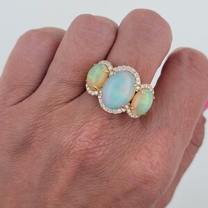 May include: A gold ring with three oval-shaped opals set in a halo of diamonds. The opals are a mix of white, green, and blue.