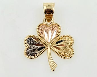 May include: A gold-toned shamrock pendant with three heart-shaped leaves. Each leaf has a different textured finish: black, gold, and rose gold. The pendant has a textured border and a loop for a chain.