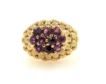 May include: A gold ring with a central cluster of deep purple gemstones. The ring's band has a textured, woven design, adding to its distinctive look. The gemstones are closely set, enhancing the ring's overall elegance and appeal.