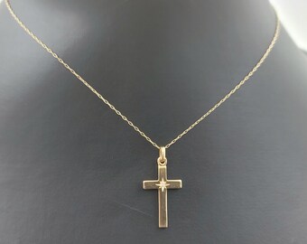 Könnte beinhalten: Goldfarbene Kreuz-Halskette mit einem kleinen diamantartigen Akzent in der Mitte des Kreuzes. Die Halskette hat eine zarte Kette und wird vor einem dunklen Hintergrund präsentiert.