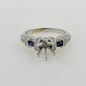 May include: A silver engagement ring setting with a center stone prong setting and two blue sapphires on either side. The band is accented with small diamonds.