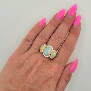 May include: A gold ring with three oval-shaped opals set in a halo of diamonds. The ring is on a hand with pink nail polish.