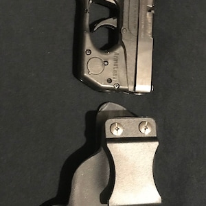 Puede incluir: Una pistola negra con corredera plateada y una funda negra. La pistola tiene el texto "Ruger" y "ArmaLaser" en ella.