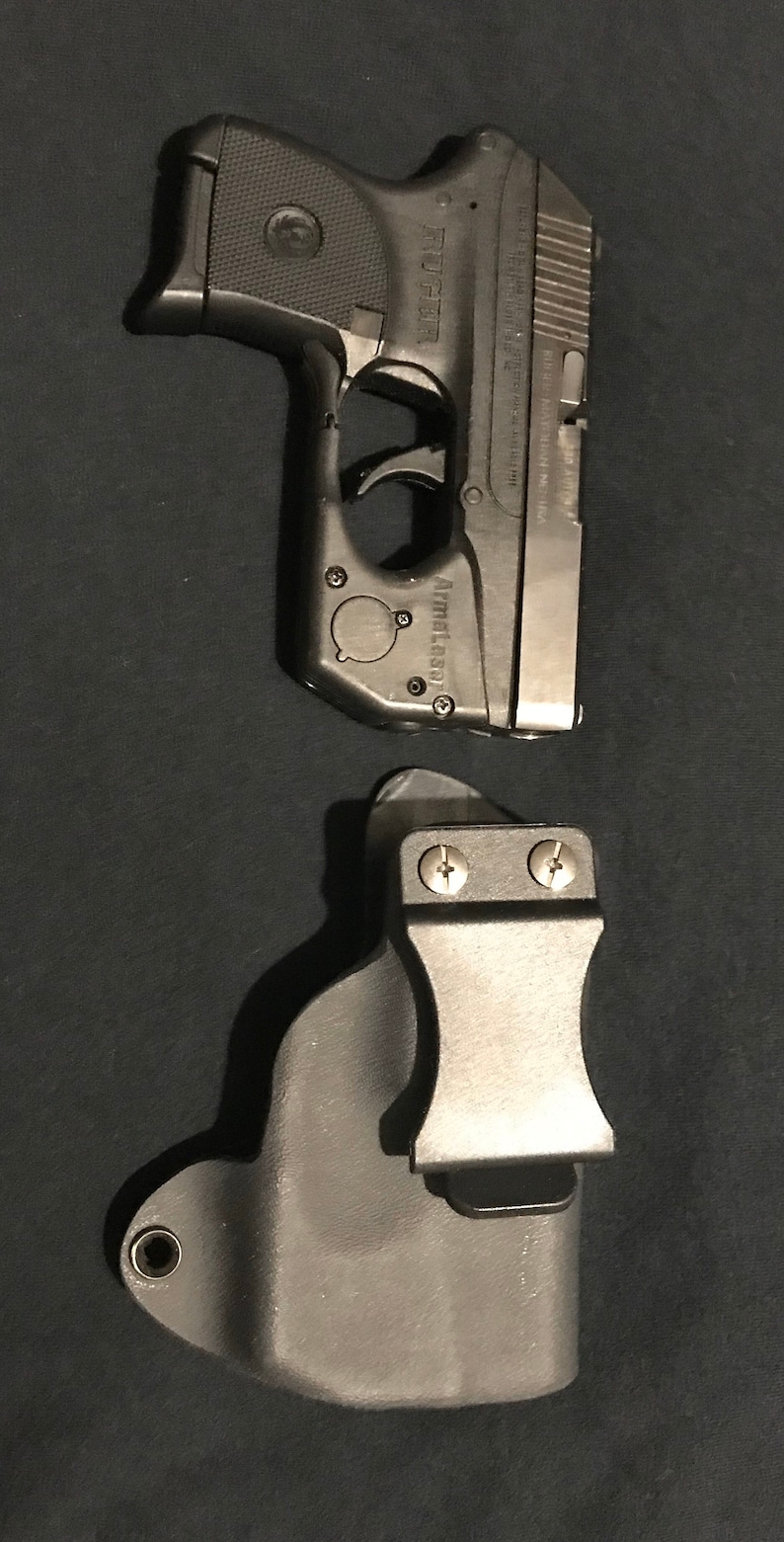 Puede incluir: Una pistola negra con corredera plateada y una funda negra. La pistola tiene el texto "Ruger" y "ArmaLaser" en ella.
