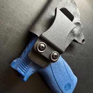 May include: A black molded holster with a textured finish and a black belt clip secured by two screws. The holster contains a bright blue firearm with a textured grip and a visible logo on the handle, showing the slide and rear sight area.