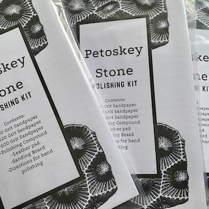 May include: Three white paper packets with black text and a black and white image of a Petoskey stone. The text on the packets reads "Petoskey Stone Polishing Kit" and lists the contents of the kit: "60 Grit Sandpaper", "220 Grit Sandpaper", "600 Grit Sandpaper", "Polishing Compound", "Leather pad", "Sanding Board", and "Directions for hand polishing".