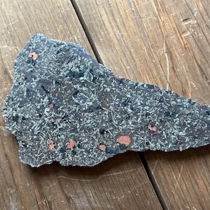 May include: A uniquely shaped, dark gray stone with a speckled pattern of green and black, and small copper-colored inclusions. The stone is irregularly shaped and rests on a wooden surface.