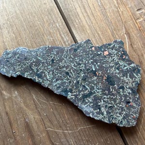 May include: A dark, irregularly shaped stone with a textured surface. The stone is primarily dark gray with patches of green and small flecks of orange. The stone is lying on a wooden surface.