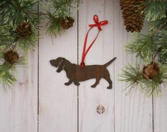 May include: A wooden Christmas ornament shaped like a dachshund dog with a red ribbon attached. The ornament is brown and has a rustic finish. It is hanging against a white wood background.