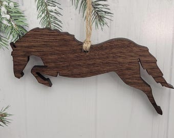 May include: A wooden horse ornament in a jumping pose, with a natural wood grain finish. The horse is suspended by a jute twine string, suitable for hanging. The ornament is designed for holiday or seasonal decor.