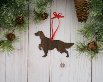 May include: A wooden Christmas ornament shaped like a brown dog with a red ribbon attached. The ornament is hanging against a white wood background with pine branches and pine cones.