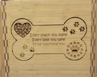 May include: A light-colored wooden cutting board with a bone-shaped design. The design includes a heart filled with paw prints, paw prints, and the text "Every snack you make, Every bite you take, I'll be watching you."