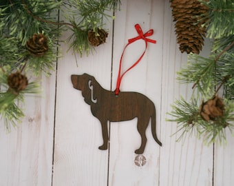May include: A wooden Christmas ornament shaped like a brown Bloodhound dog with a red ribbon attached. The ornament is hanging against a white wood background.