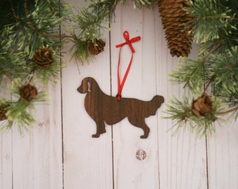 May include: Wooden Christmas ornament shaped like a golden retriever dog with a red ribbon attached. The ornament is hanging against a white wood background.