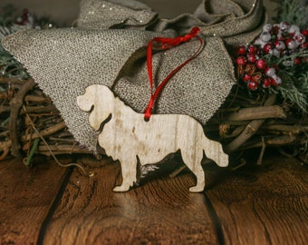 May include: A wooden dog ornament in the shape of a spaniel, with a red ribbon attached. The ornament is sitting on a wooden surface with a burlap background.