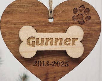 May include: A wooden heart-shaped ornament with a light-colored bone-shaped cutout that reads "Gunner". The ornament has a paw print and the dates "2013-2025" engraved on it. A twine loop is attached for hanging.