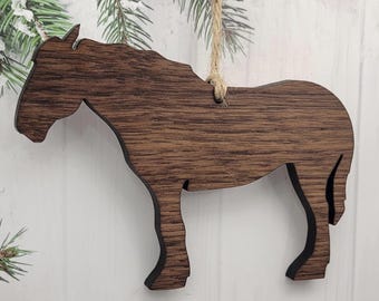 May include: A wooden horse ornament with a dark brown finish. The horse is cut out in a side profile, with a natural wood grain visible. A twine loop is attached for hanging. The ornament is a rustic decoration.