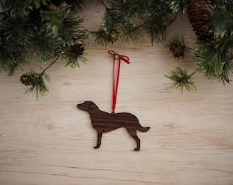 May include: A wooden dog ornament with a red ribbon for hanging. The ornament is a silhouette of a dog, crafted from dark wood. The background is a light wood surface with pine branches and pine cones.