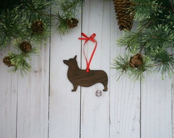 May include: Wooden Christmas ornament shaped like a corgi dog silhouette with a red ribbon. The ornament is hanging against a white wood background.