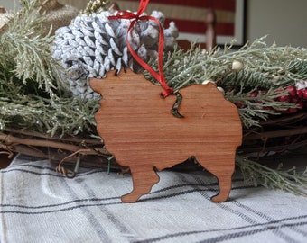 May include: A wooden Christmas ornament shaped like a Pomeranian dog. The ornament is a light brown color and has a red ribbon attached to the top.
