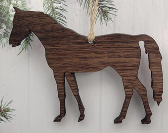 May include: A wooden horse ornament with a natural wood grain finish. The horse is depicted in a side profile, with a jute string for hanging. The ornament is a decorative item, suitable for holiday or rustic decor.