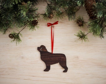 May include: A wooden ornament shaped like a Newfoundlander dog, with a red ribbon attached. The ornament is hanging from a pine branch on a wood background.